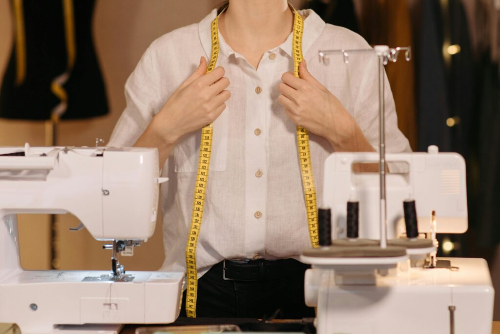 Tailor in a white shirt uses a measuring tape with sewing machines in a workspace.